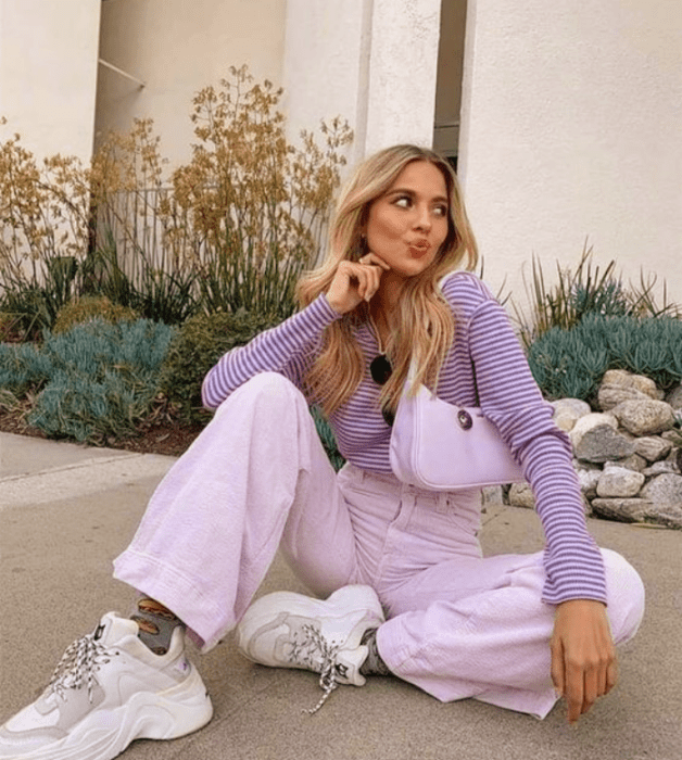 chica rubia con cárdigan lila, pantalones lila morado pastel, tenis blancos y bolso de cuero pequeño color lila