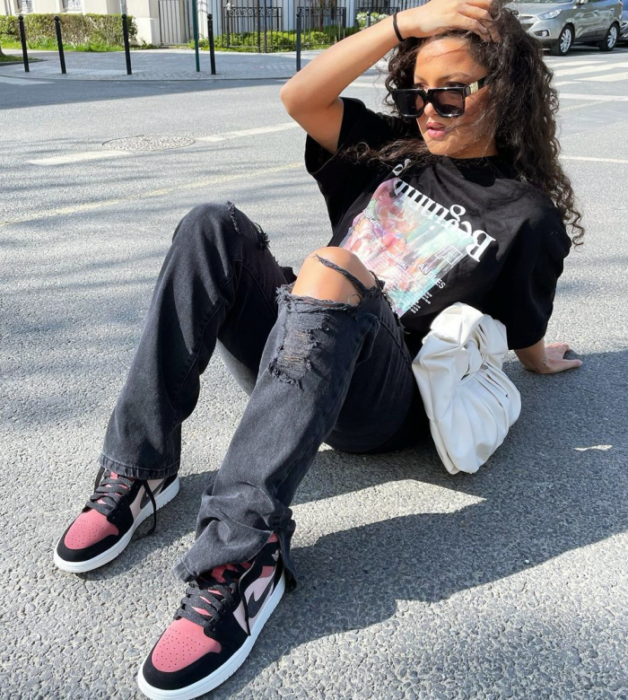 chica morena de cabello chino usando una camiseta oversized negra gráfica, jeans negros holgados rotos, tenis blancos con negro y rojo y bolso blanco