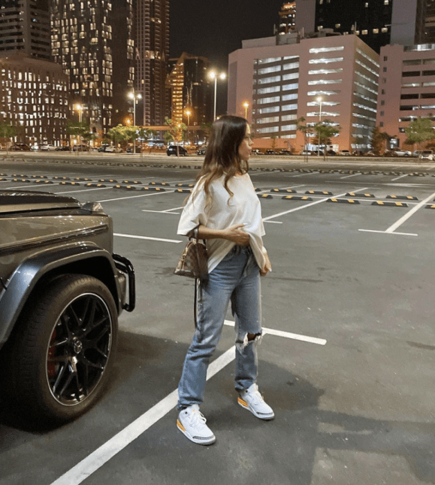 chica de cabello castaño usando una camiseta blanca oversized, jeans holgados rotos, tenis blancos, bolso café de cuero