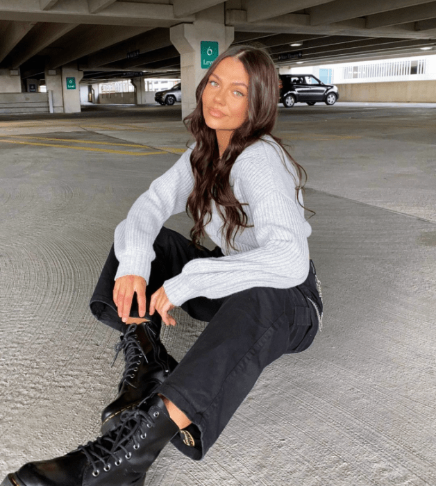 chica de cabello castaño usando una sudadera blanca gris, jeans negros rectos, botines negros de cuero