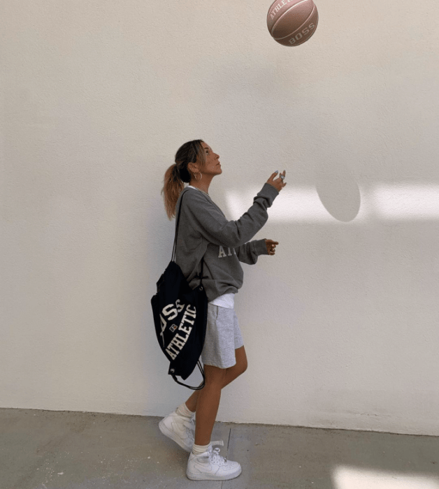 chica de cabello claro usando una sudadera gris oversized, shorts grises claros, tenis blancos, bolso tote bag negro con letras blancas