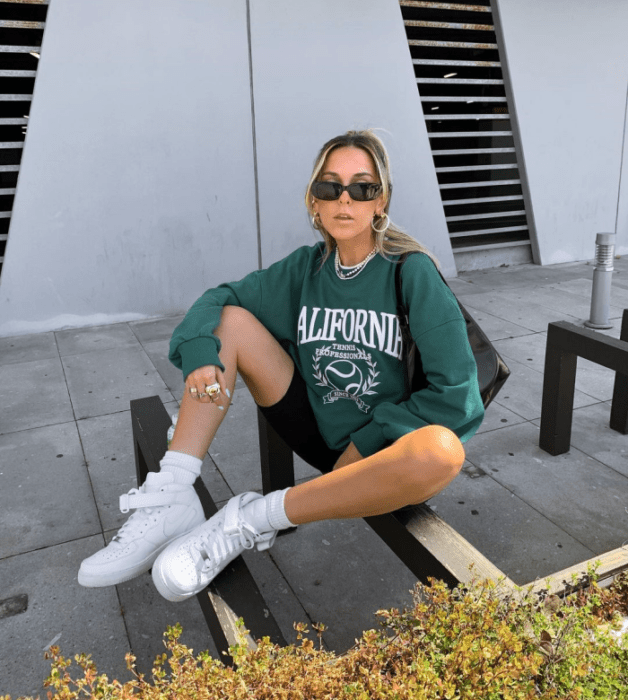 chica rubia con lentes de sol, sudadera verde oversized, tenis blancos y shorts de ciclismo negros