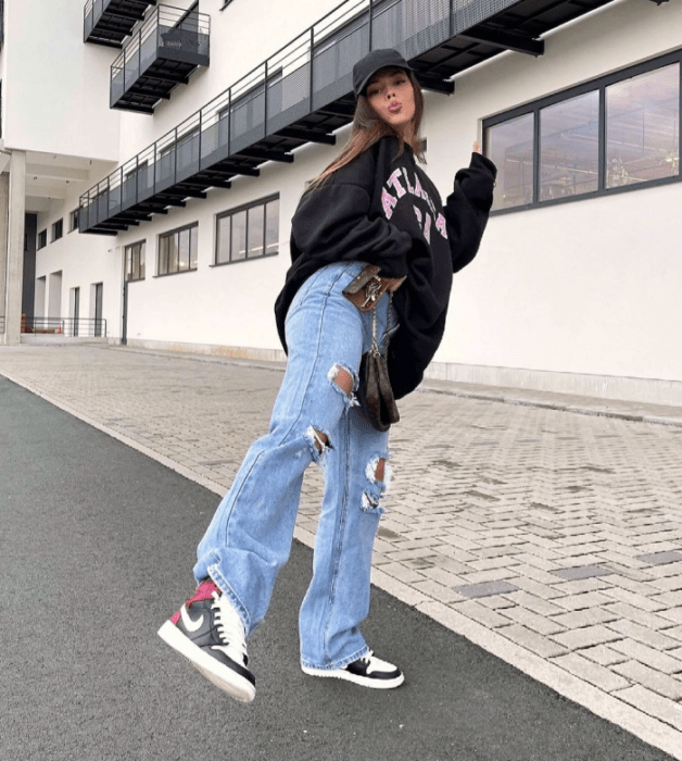 chica de cabello claro usando una gorra negra, sudadera oversized negra, jeans holgados rotos, tenis blancos con negro