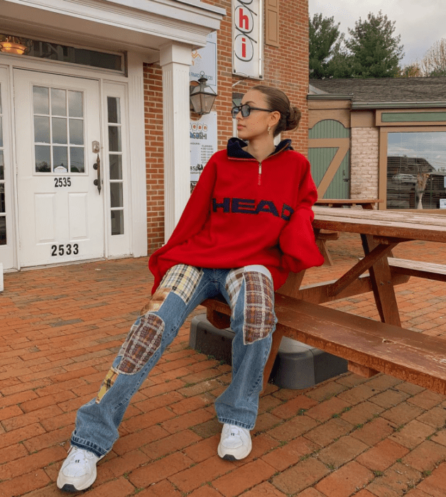 chica de cabello castaño usando lentes de sol, sudadera oversized roja de cuello azul marino, jeans holgados de mezclilla con cuadros rojos, tenis blancos