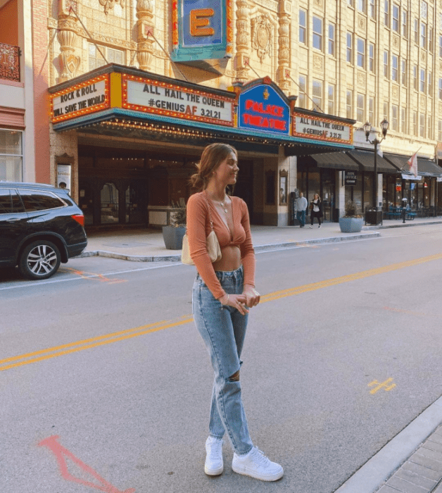 chica de cabello castaño usando un crop top rosa de manga larga, jeans rotos ajustados, tenis blancos y bolso beige pequeño