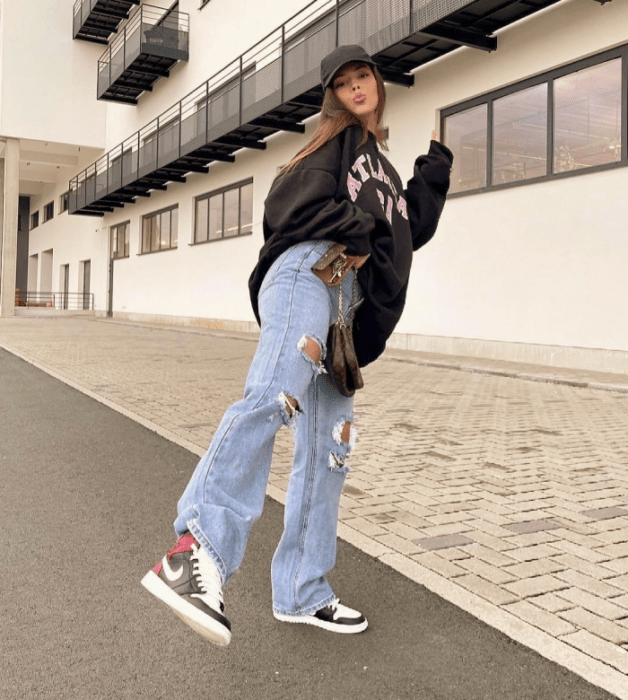 chica de cabello castaño con sudadera negra overszied, jeans rotos holgados, tenis blancos con negro y bucket hat negro