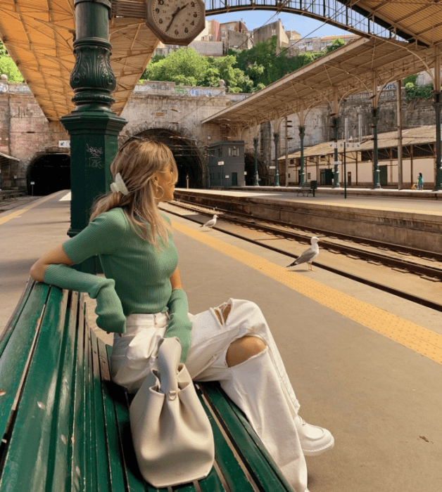 chica rubia con top verde tejido de manga larga, jeans blancos rotos, tenis blancos, bolso blanco grande cuero