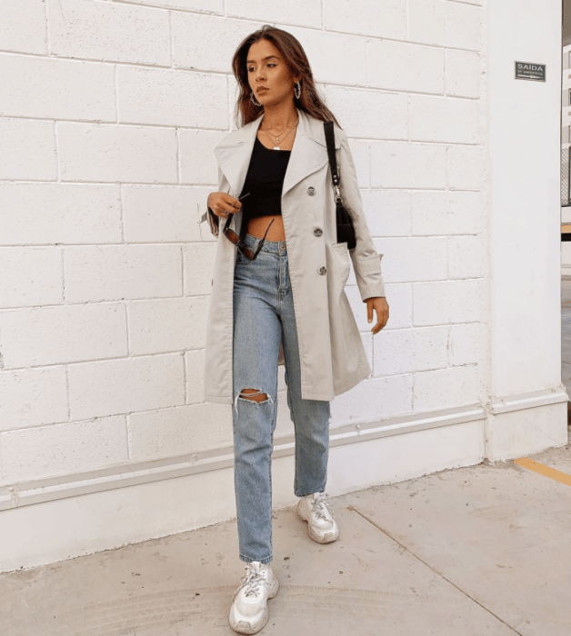 chica de cabello castaño usando un crop top negro, abrigo largo beige, jeans ajustados rotos, tenis blancos, bolso negro de cuero