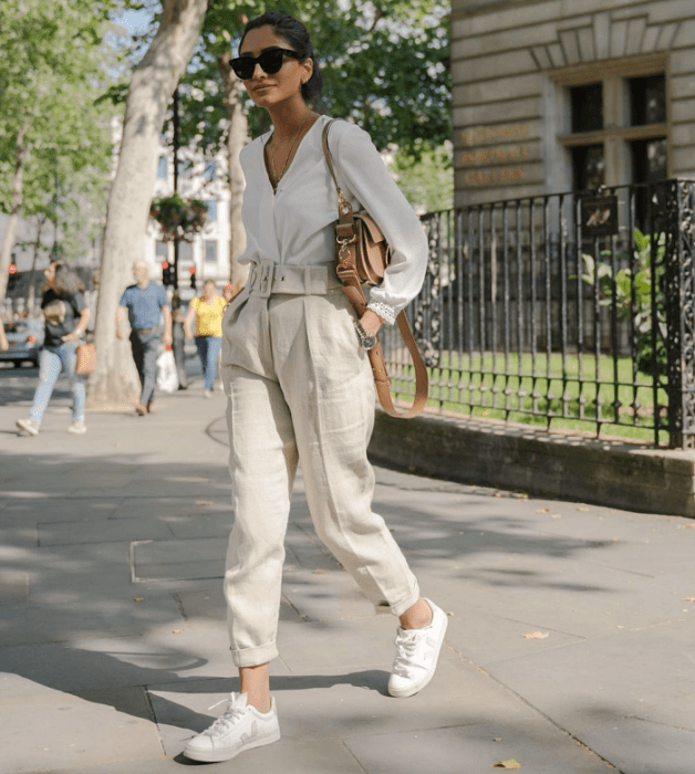 chica morena con lentes de sol, cárdigan de manga larga de botones, pantalones blancos de vestir, tenis blancos, bolso café de mano