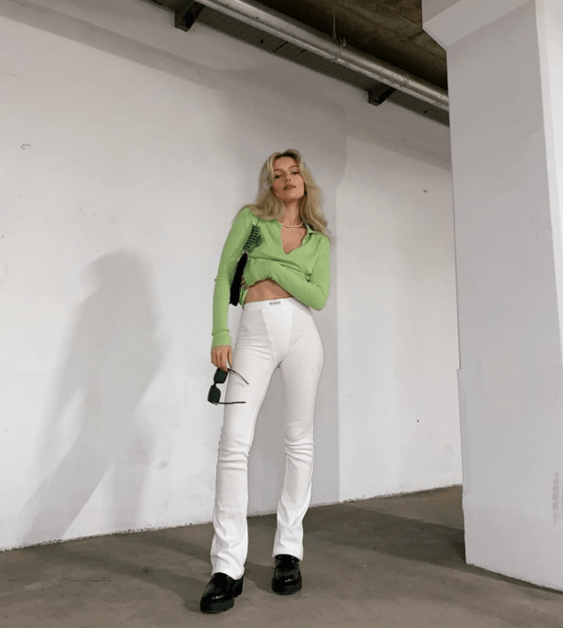 chica rubia con top verde claro de manga larga, leggings blancos ajustados, botas negras de cuero, bolso pequeño negro