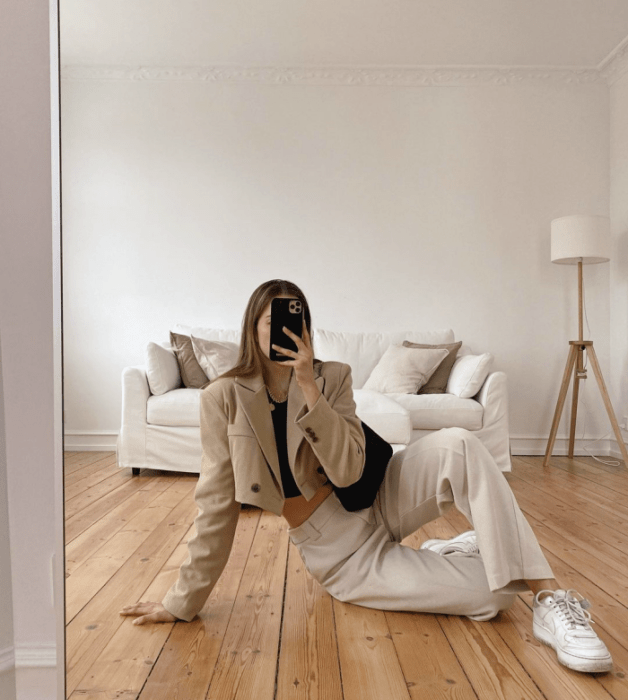 chica de cabello claro usando un top negro, blazer corto beige, pantalones blancos, tenis blancos, bolso negro de cuero