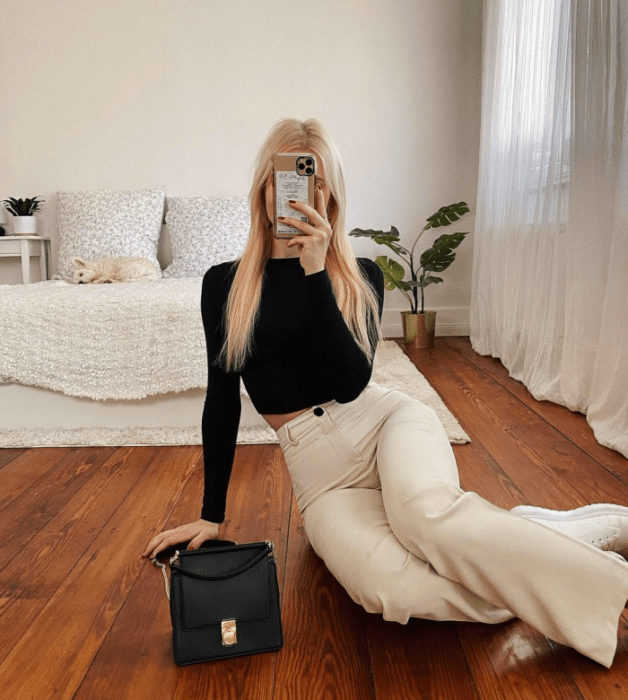 chica rubia con top negro de manga larga, pantalones blancos acampanados, bolso pequeño negro de cuero