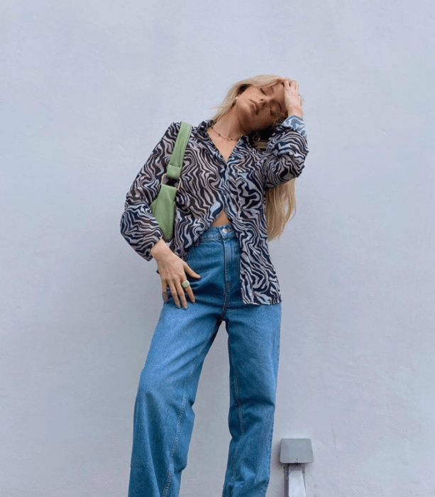 chica rubia con camisa de vestir semitransparente negra con blanco, jeans holgados, bolso verde de cuero pequeño