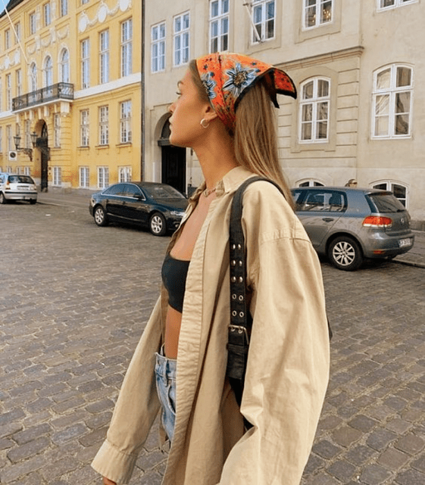 chica de cabello rubio largo usando una mascada en el cabello, crop top de tubo negro, camisa beige oversized, jeans ajustados azules