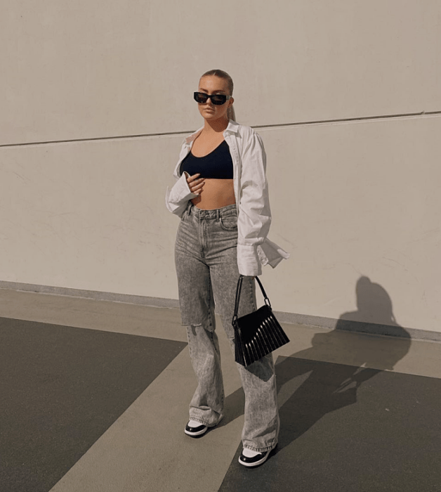 chica rubia con lentes de sol, crop top negro de tirantes, camisa blanca de botones, jeans grises holgados, tenis blancos con negro, bolso pequeño negro