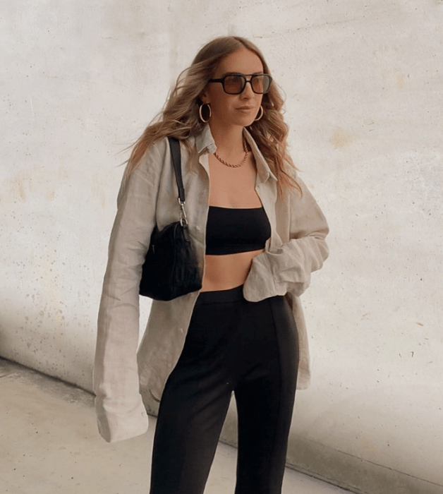 chica rubia con lentes de sol, crop top negro, camisa de vestir beige, pantalones negros ajustados a la cintura y bolso negro de cuero pequeño