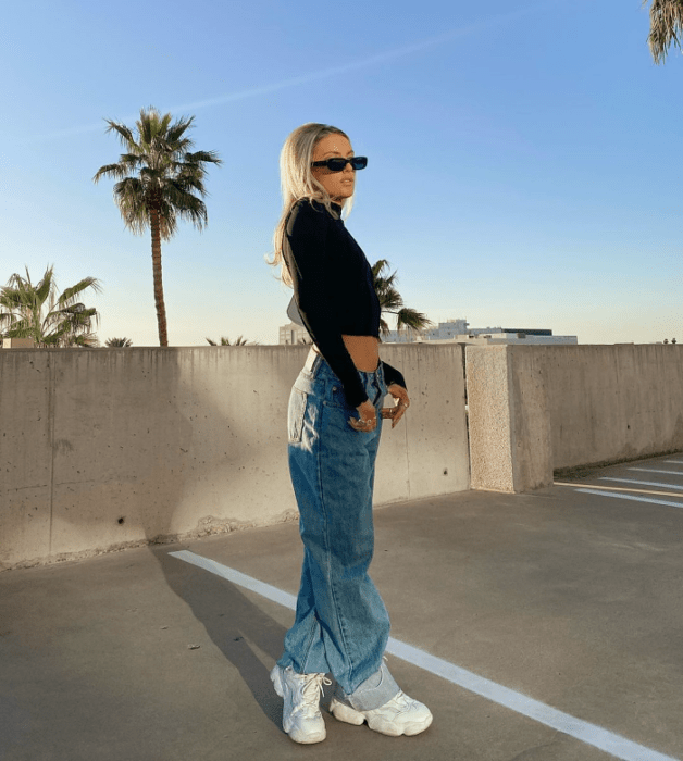 chica rubia con lentes de sol, crop top negro de manga larga, jeans holgados a la cintura, tenis blancos deportivos