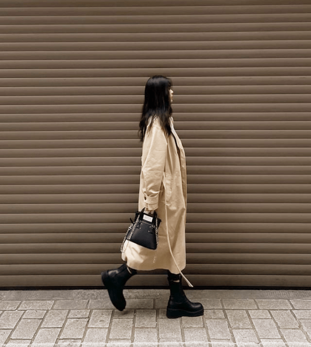 chica de cabello castaño largo usando un abrigo largo beige, botines negros de cuero, bolso negro de cuero