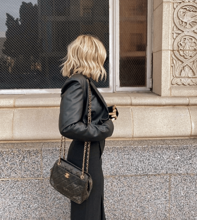 chica rubia de cabello corto usando una chaqueta de cuero, pantalones negros y bolso negro de mano