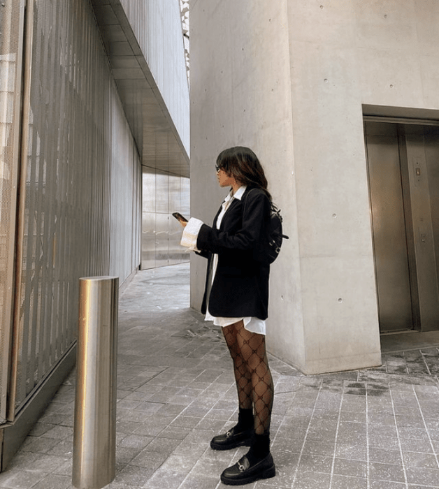 chica de cabello castaño usando una camisa blanca de vestir, blazer negro oversized, medias negras de red, botines negros de cuero