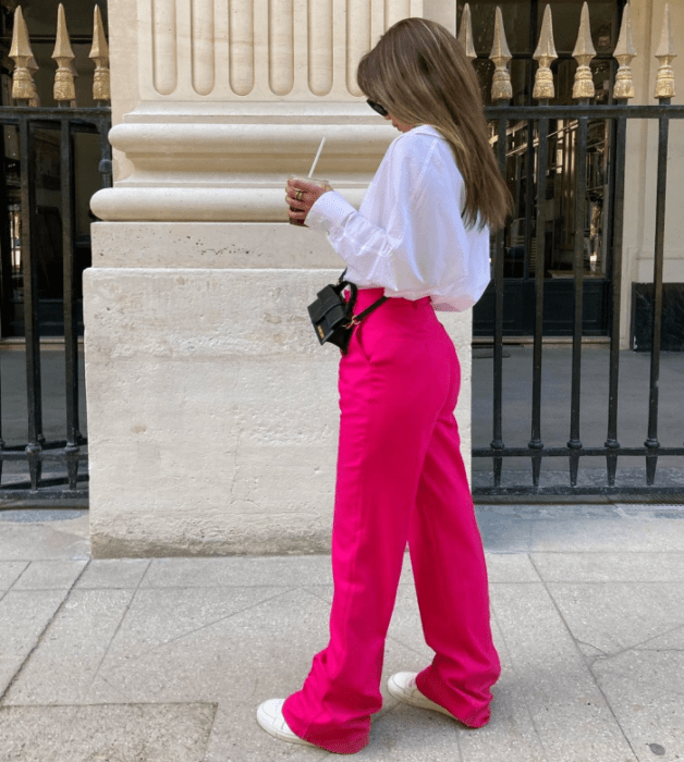 chica de cabello claro usando una camisa de vestir blanca, pantalones rosas fucsias, tenis blancos, bolso pequeño negro de cuero