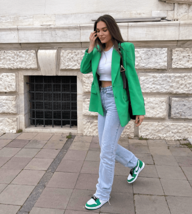 chica de cabello castaño usando un crop top blanco, blazer verde brillante, jeans ajustados celestes, tenis blancos con verde y negro, bolso negro