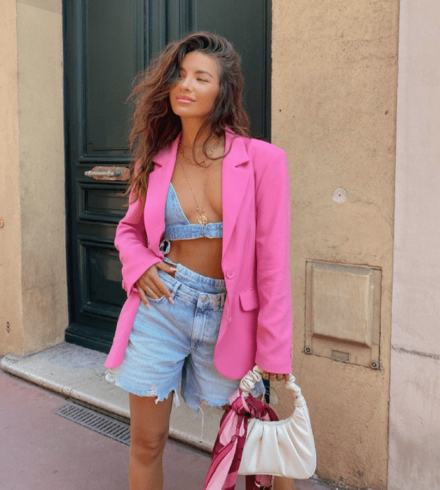 chica morena de cabello largo usando un bralette de mezclilla, shorts de mezclilla, blazer rosa fucsia, bolso pequeño beige de cuero