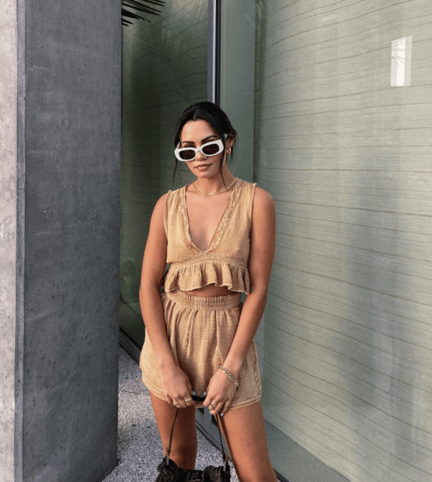 chica de cabello castaño usando lentes de sol blancos, crop top beige de tirantes, shorts beige, bolso negro de cuero