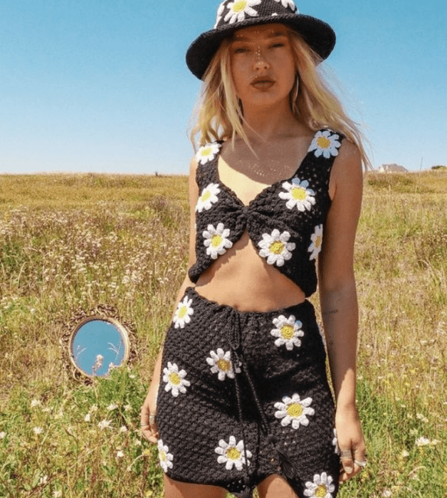 chica rubia con bucket hat negro tejido con margaritas blancas, crop top negro de flores blancas, minifalda negra tejida con flores blancas margaritas