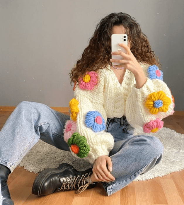 chica de cabello chino usando un cárdigan beige con flores grandes rosas, azules, amarillas y verdes, jeans holgados y botines negros de cuero