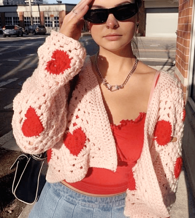 chica con lentes de sol, top rojo tejido, cárdigan rosa claro con corazones rojos, jeans ajustados