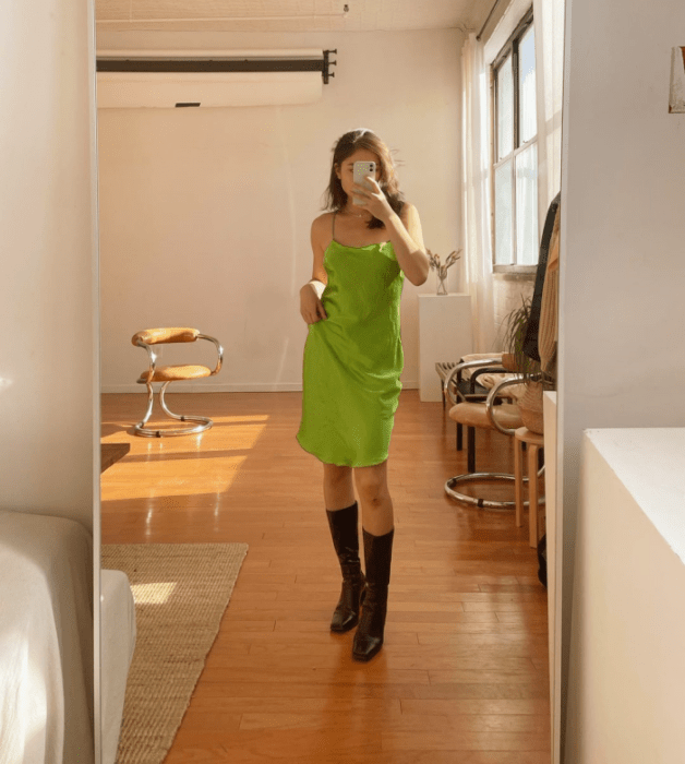 chica de cabello castaño usando un vestido de satén verde de tirantes con botas largas negras de cuero