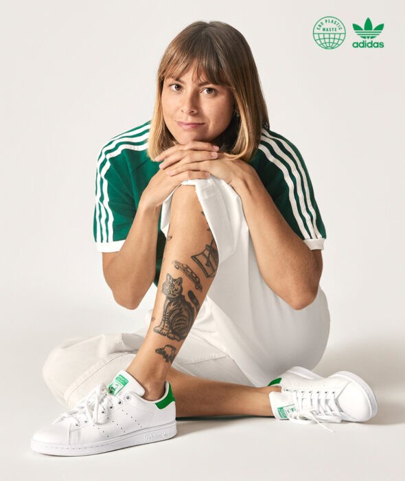 Linea de ropa y calzado de Adidas, Stan Smith Disney; sudadera o hoodie de cuello redondo, con gorro, color hueso y estampado verde de la rana René; mujer sentada en el suelo, sonriendo y con las manos en las rodillas, cabello rubio castaño corto a los hombros y con fleco, tatuaje de gato en la pierna