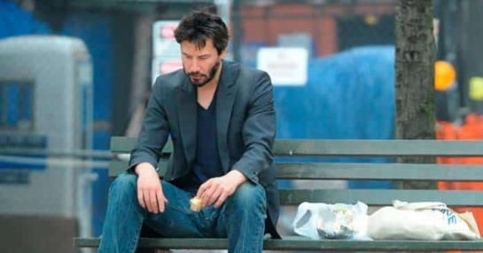 Keanu Reeves eating a sandwich sitting in a park 