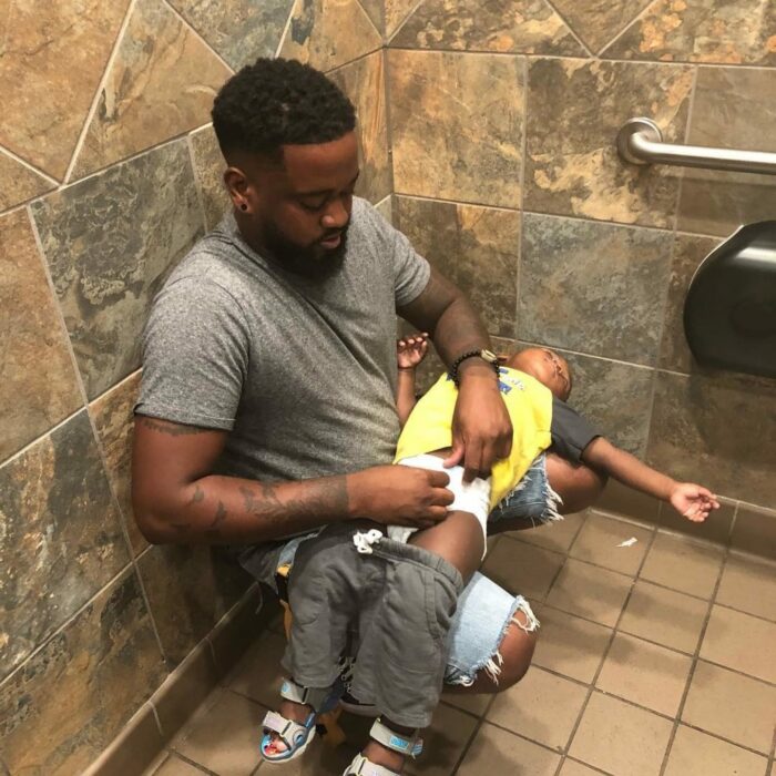 foto de un hombre cambiando el pañal a su hijo sentado en el piso de un baño público 