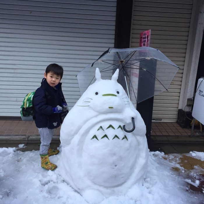 niño parado a un lado de una escultura de nieve en forma del personaje Totoro