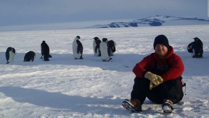 Cole Heinz sentado en la nieve con pingüinos a sus espaldas