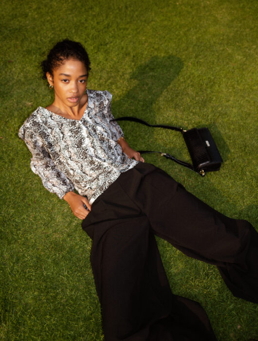 Brunette woman lying on the grass in the garden wearing an animal print blouse, baggy pants and a black bag