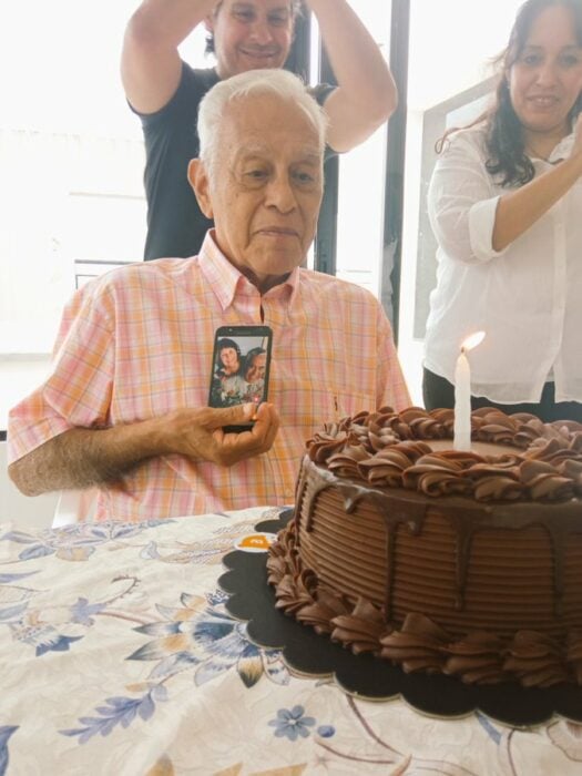 Abuelit sosteniendo un teléfono celular; Al soplar las velas del pastel, abuelo saca foto de su esposa; es su primer cumple sin ella