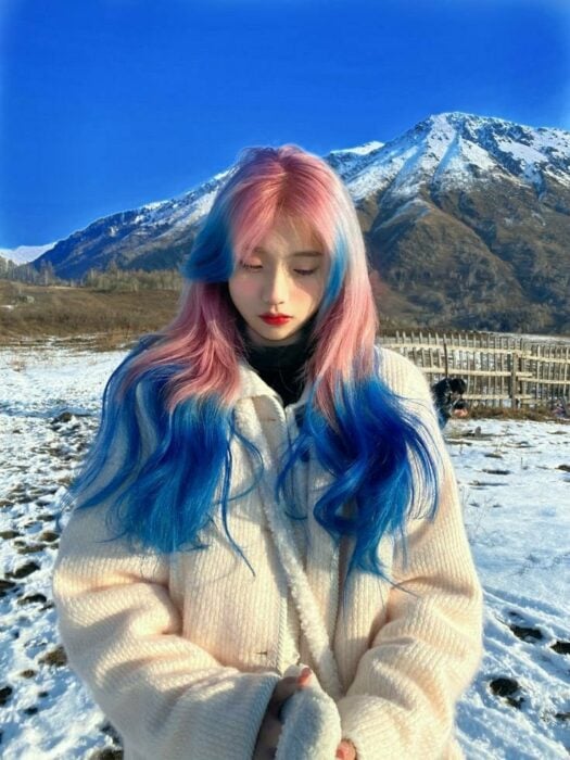 chica asiática en medio de unas montañas nubladas mostrando su cabello en color rosa con azul 