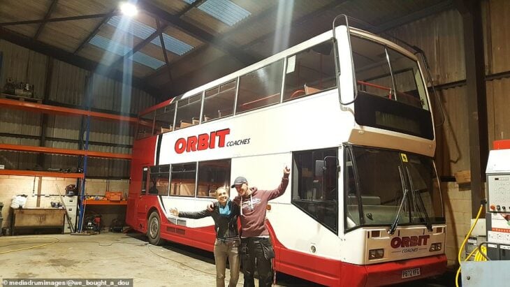 photograph of a woman and a man in front of a double-decker bus in a warehouse 