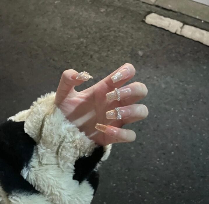 mano de una mujer mostrando uñas color nude con brillitos y detalles de moño en el dedo anular 