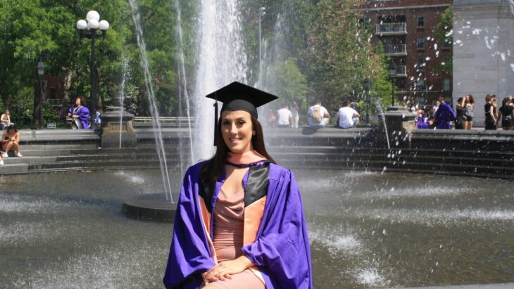 Foto de una chica¿ con birrete y toga sentada frente a una fuente de agua 