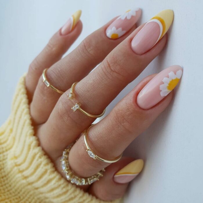 mano de una mujer mostrando sus anillos y uñas en color blanco con amarillo con diseños de flores 