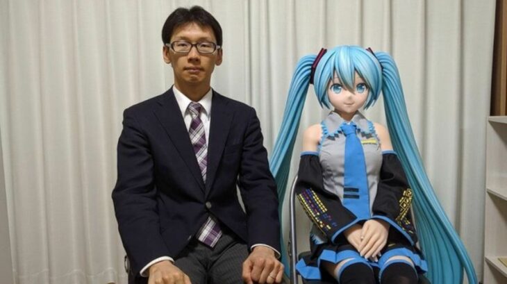 Akihiko Kondo sentado a lado de una muñeca de Miku, un personaje ficticio 