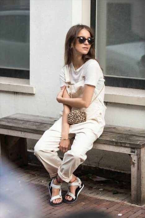 girl wearing white overalls with sunglasses sitting on a bench in a street 