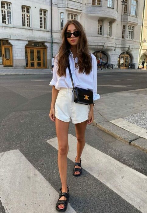 woman posing across the street wearing a denim shorts with a white blouse with sports sandals 