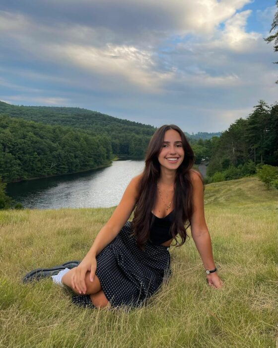 Fotografía de una chica sentada sobre un pasto verde frente a un lago 