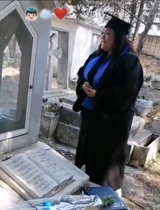 Chica parada con su toga y birrete frente a la tumba de su padre