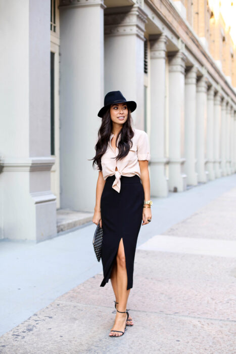 photo of a woman walking down a street wearing a skirt with a slit in the front and a fedora 